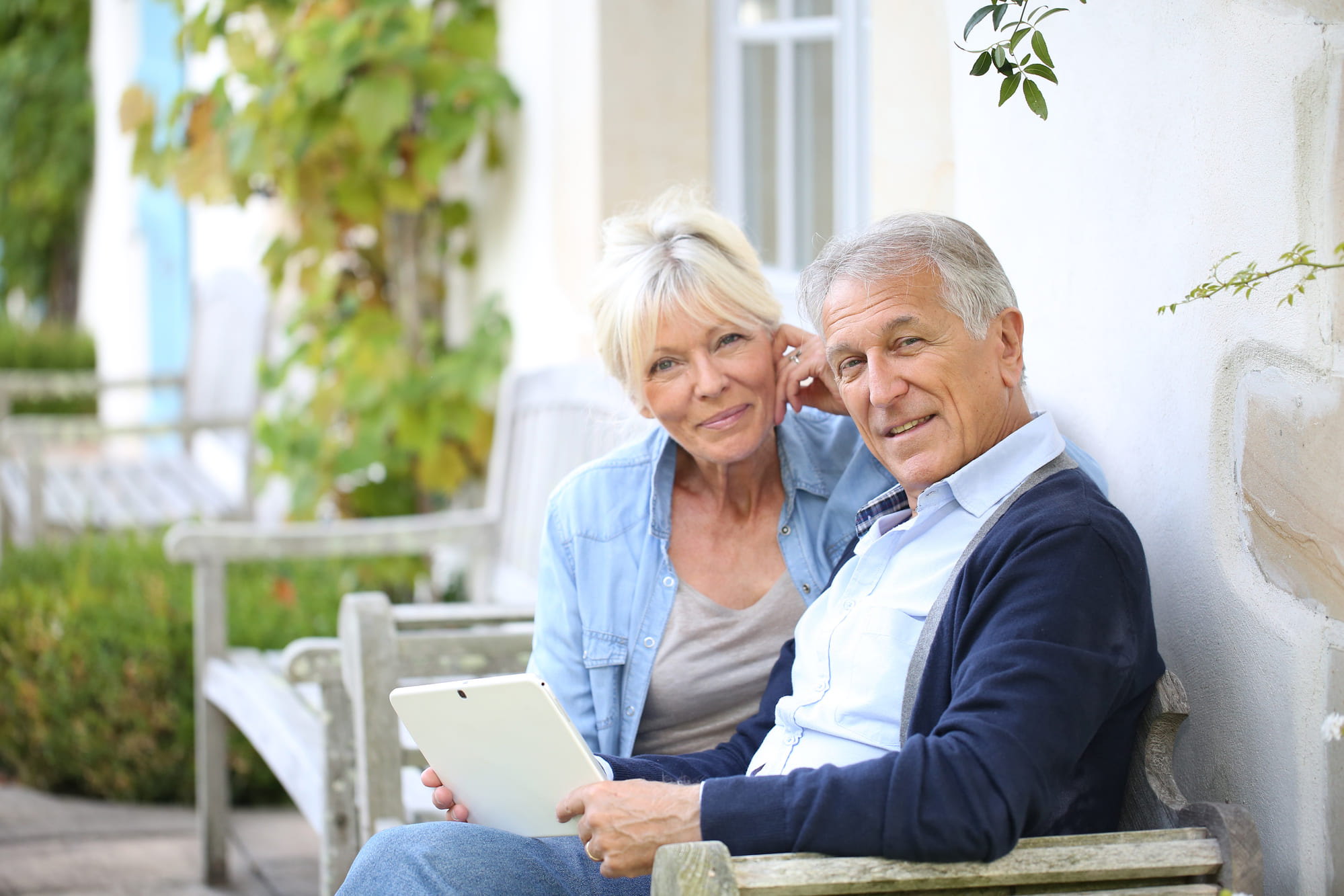 Zwei lächelnde Senioren beim Surfen im Internet auf der Bank vor einem Haus | Wohnen im Alter